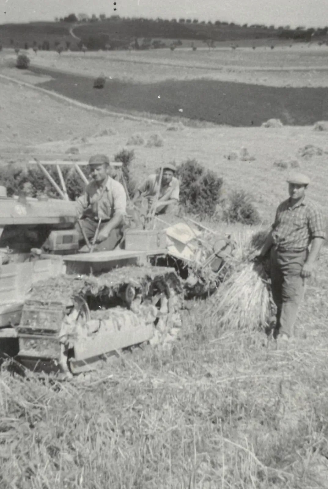 Una foto d’epoca: uomini al lavoro nel campo mietuto, accanto a una macchina cingolata, con le colline sullo sfondo.