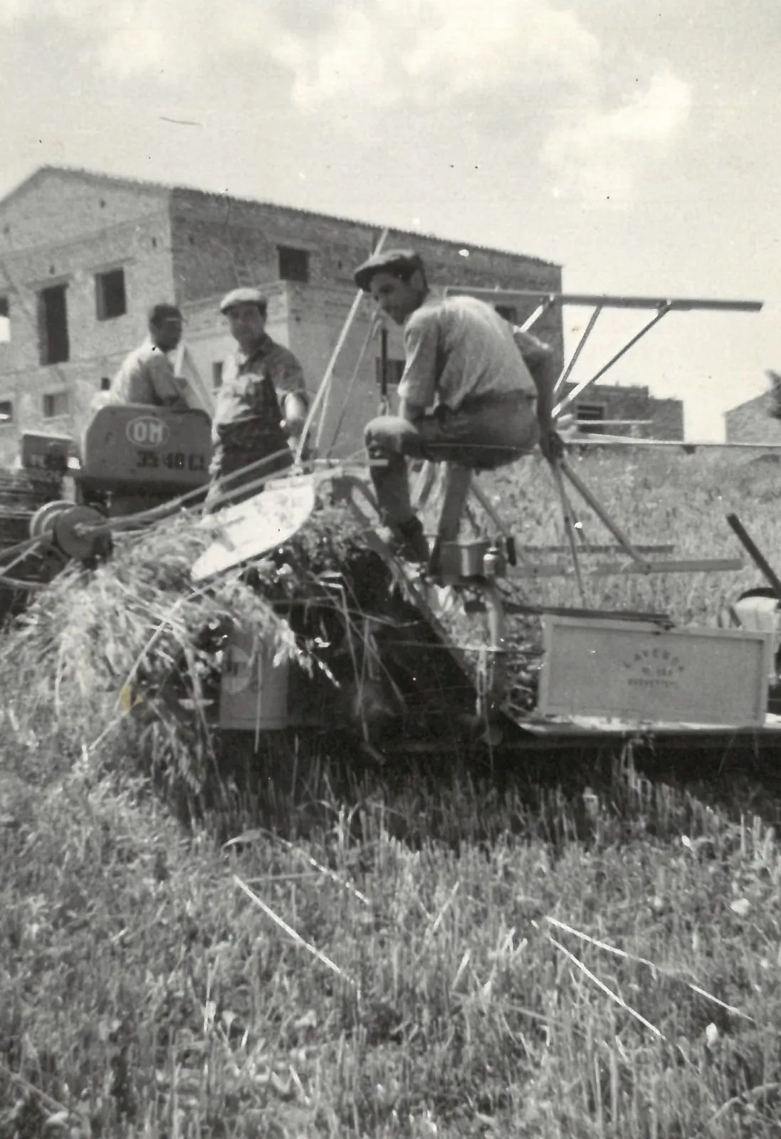 Una foto d’epoca: uomini su una macchina agricola nel campo, davanti a una casa in costruzione.